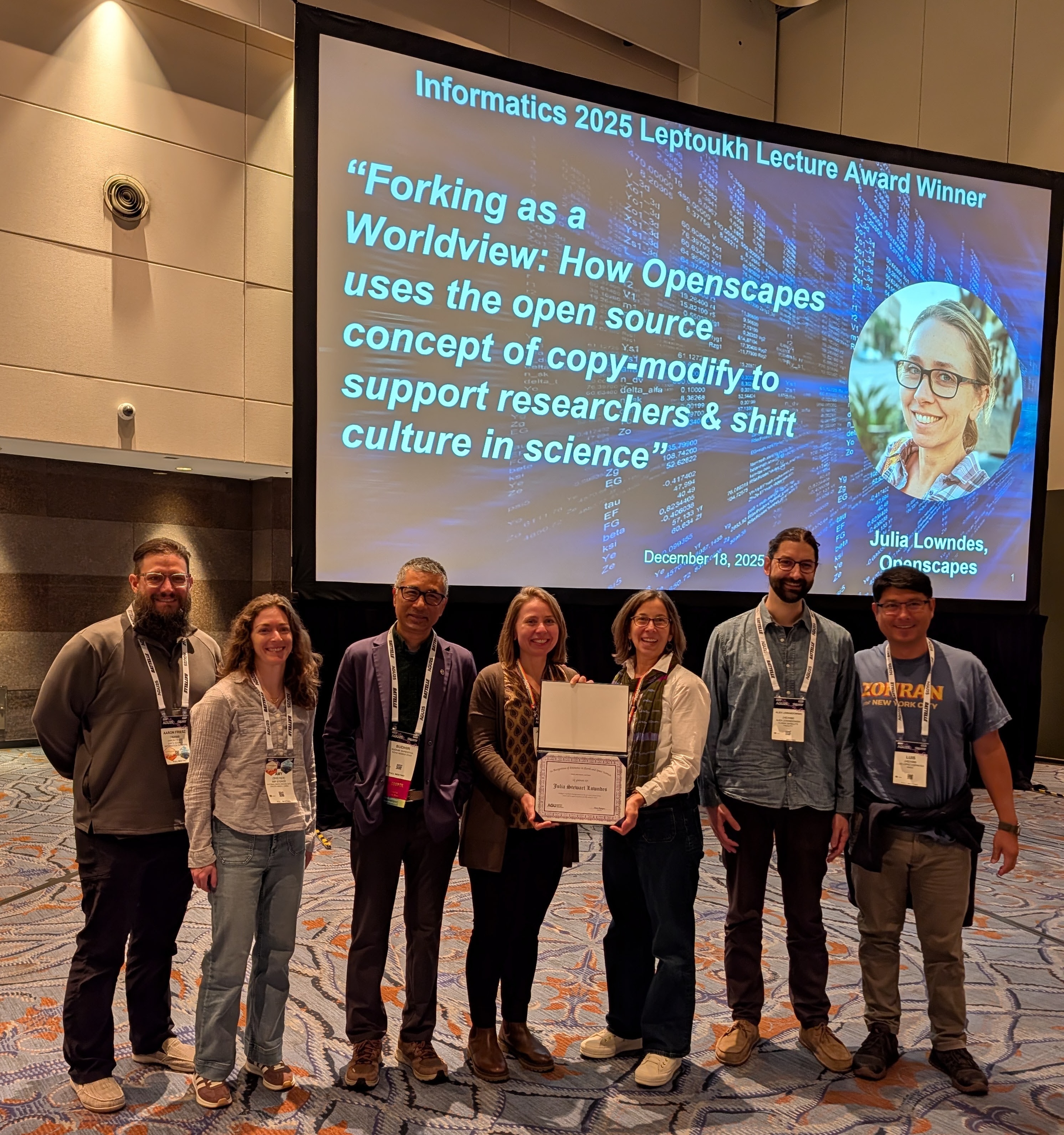 6 women and men smiling, wearing conference lanyards, standing in front of a large screen projecting a headshot of Julie Lowndes and her talk title Forking as a worldview: How Openscapes uses the open source concept of copy-modify to support researchers & shift culture in science