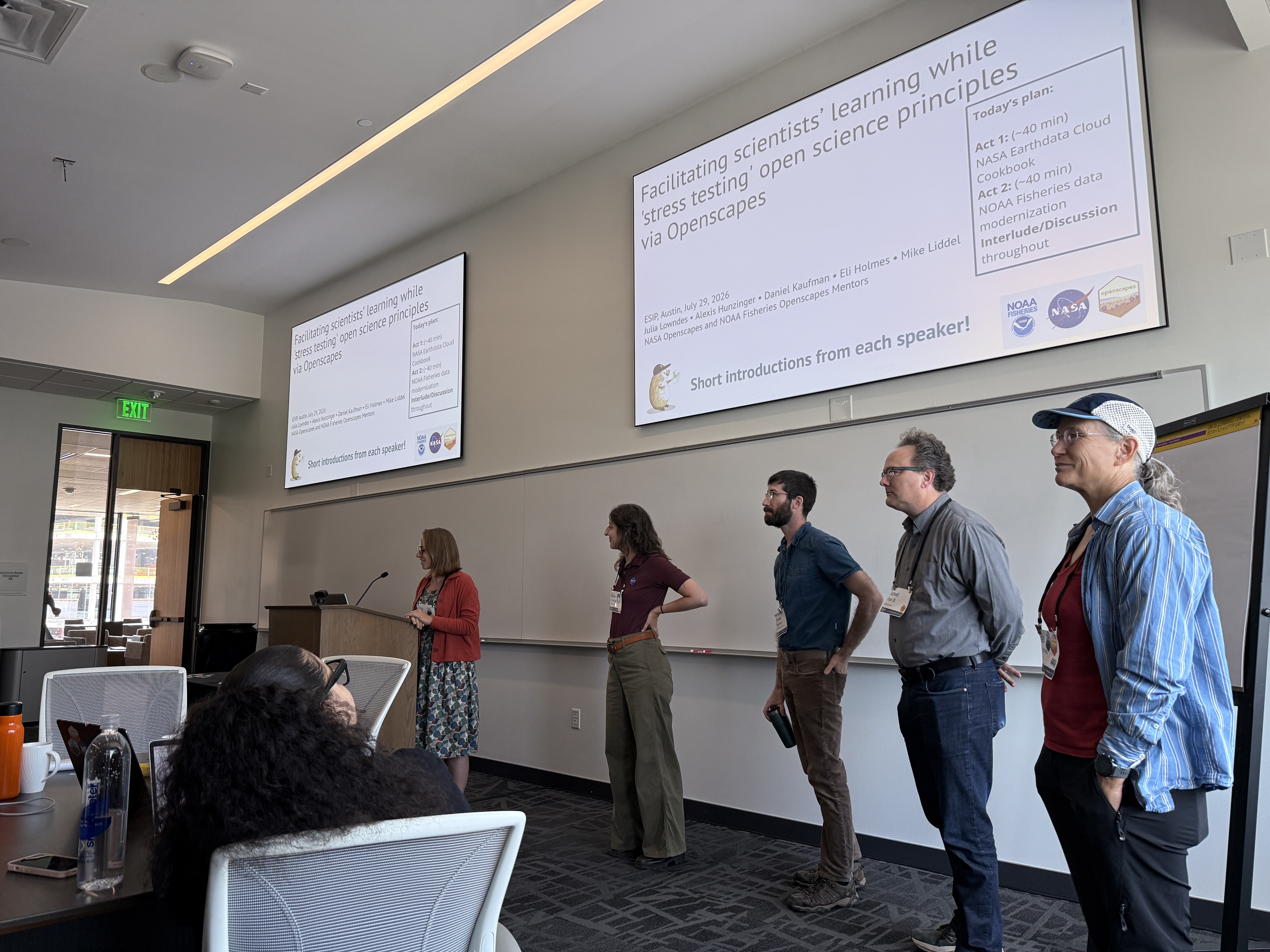 Presenters standing at the front of a conference room. Behind them, projected slides introduce a session on facilitating scientists’ learning while stress-testing open-science principles through Openscapes.
